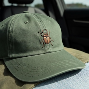 Puede incluir: Gorra de béisbol verde oliva con un diseño de escarabajo bordado en marrón en la parte delantera. La gorra tiene una visera curva y un cierre de correa de tela. Accesorio de moda.
