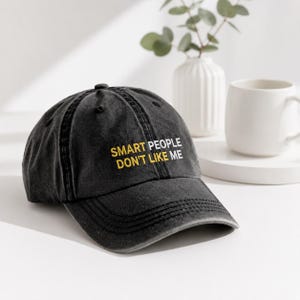 Puede incluir: Una gorra de b&eacute;isbol gris carb&oacute;n con acabado desgastado que presenta el texto bordado "SMART PEOPLE" en amarillo y "DON'T LIKE ME" en blanco en el panel frontal. La gorra tiene una visera curva y una corona estructurada con un bot&oacute;n.