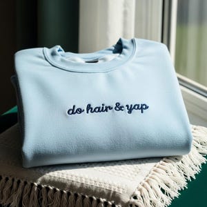 May include: A pale blue sweatshirt featuring the embroidered phrase "do hair & yap" in navy blue. The garment is neatly folded on a cream-coloured blanket with a fringed edge. The image highlights the soft texture and colour of the sweatshirt.