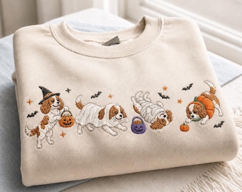 Può includere: Una felpa girocollo beige con un ricamo raffigurante quattro cani Cavalier King Charles Spaniel in costume di Halloween. I cani sono vestiti da strega, fantasma, mummia e zucca, circondati da piccoli pipistrelli e stelle di colore arancione.