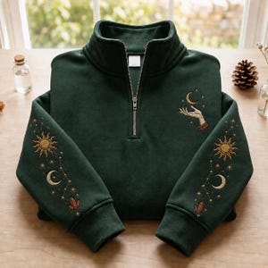 May include: A dark green quarter-zip sweatshirt featuring intricate embroidery on both sleeves and the chest. The sleeve designs include a sun, moon, stars, and crystals. The chest embroidery depicts a hand reaching toward a crescent moon.