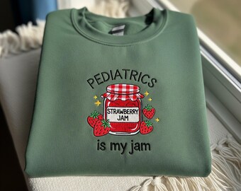 May include: Green sweatshirt with embroidered text and graphic. The design features the word "PEDIATRICS" above a jar of strawberry jam, surrounded by strawberries and the phrase "is my jam".