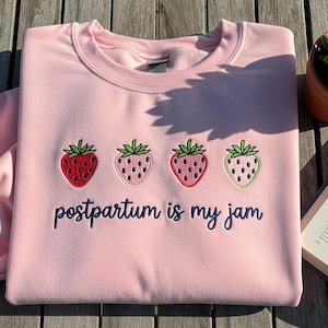 以下が含まれることがあります： 4つのイチゴと「postpartum is my jam」というフレーズがネイビーブルーで刺繍された、明るいピンク色のスウェットシャツ。 イチゴは赤、ピンク、緑の色合いです。 スウェットシャツは柔らかい素材で作られています。