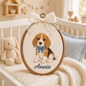 Puede incluir: Un bastidor de bordado ovalado de madera que contiene un panel de tela con un cachorro de beagle bordado con una pajarita azul. El nombre Annie está bordado en letra azul debajo del cachorro. Un lazo de encaje color crema está sujeto arriba.