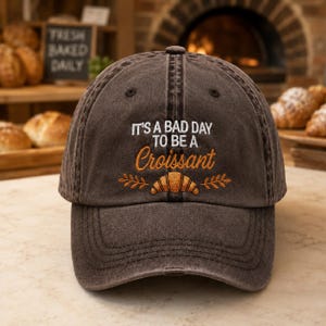 Könnte beinhalten: Eine anthrazitfarbene Baseballkappe mit Used-Look und weiß-orangener Stickerei mit der Aufschrift IT'S A BAD DAY TO BE A Croissant, ergänzt durch eine kleine Grafik eines Croissants, das von zwei Weizenähren auf der Vorderseite flankiert wird.
