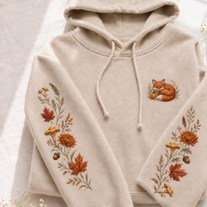May include: A beige hooded sweatshirt featuring embroidered designs on the sleeves and chest. The chest displays a small fox curled around a pumpkin. The sleeves feature botanical embroidery including orange maple leaves, mushrooms, and acorns.