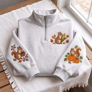 Könnte beinhalten: Ein hellgraues Sweatshirt mit kurzem Reißverschluss und Stickereien von braunen Streifenhörnchen. Ein Ärmel zeigt ein Streifenhörnchen mit einem Beerenkorb, der andere mit Kürbissen und auf der Brust ist ein Streifenhörnchen mit Mais.