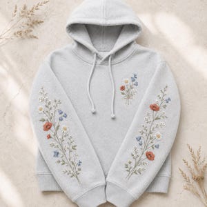 May include: A light gray hooded sweatshirt featuring delicate floral embroidery on both sleeves and the upper chest. The design includes small red poppies, white daisies, and blue bell-shaped flowers with green stems, complemented by matching drawstrings.