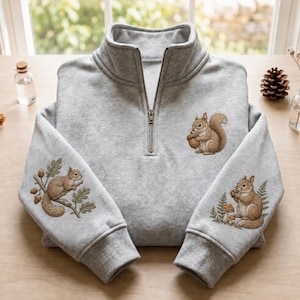 Puede incluir: Una sudadera gris claro con cremallera de un cuarto y bordados detallados de ardillas marrones. Una ardilla en el pecho sostiene una bellota, mientras que las mangas muestran ardillas sobre ramas de roble y junto a helechos y setas.