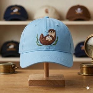 Op de afbeelding: Lichtblauwe baseballcap met een geborduurd otterontwerp. De otter is bruin en wit, drijvend in water met groene zeewier. Verschillende andere baseballcaps zijn zichtbaar op een plank op de achtergrond.