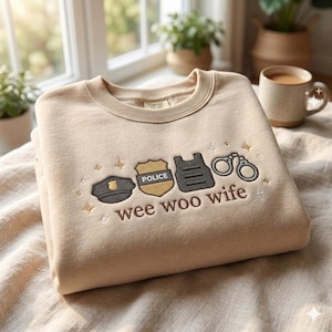 Puede incluir: Sudadera beige con bordados temáticos de la policía: gorra, placa, chaleco y esposas. El texto "wee woo wife" está bordado debajo. La sudadera está doblada y colocada sobre una superficie clara.