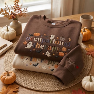 Puede incluir: Una sudadera de cuello redondo marrón oscuro con el texto bordado occupational therapy, rodeado de pequeños fantasmas, calabazas, murciélagos, estrellas y una telaraña. Debajo hay una sudadera beige con el nombre Taylor bordado en la manga.