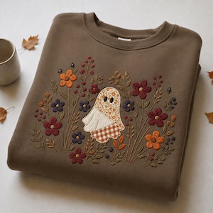 May include: A brown crewneck sweatshirt featuring an embroidered design of a ghost with a floral and gingham pattern, surrounded by various embroidered flowers and foliage in shades of orange, dark red, and purple on the front of the garment.