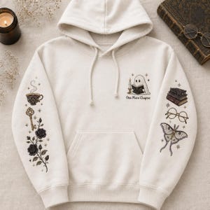 May include: A cream-colored hooded sweatshirt featuring a graphic of a ghost reading a book with the text One More Chapter. The sleeves are decorated with illustrations of a steaming teacup, a key, roses, a stack of books, glasses, and a moth.