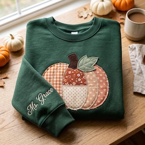 Op de afbeelding: Een donkergroene sweater met ronde hals en een grote, centrale pompoenapplicatie gemaakt van diverse stoffen in oranje, witte en bruine patronen. Op de mouw is de naam Ms. Grace geborduurd in een wit cursief lettertype.