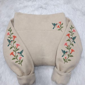 May include: A beige sweatshirt featuring detailed embroidery of hummingbirds and orange trumpet-shaped flowers. The embroidery is positioned on both sleeves and the upper right chest area, showcasing green leaves and colorful bird plumage.