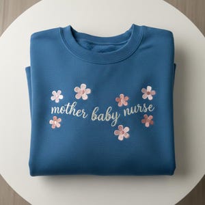 May include: A folded, teal sweatshirt featuring the embroidered words "mother baby nurse" with pink and white floral accents. The garment is made of a soft fabric and is neatly folded on a white surface.