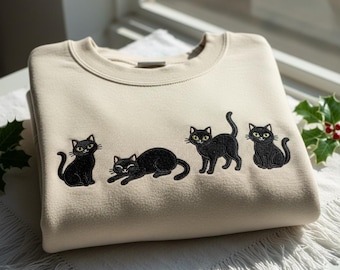 May include: A cream-coloured sweatshirt with an embroidered design of four black cats. Each cat has yellow eyes and is in a different pose. The sweatshirt is made of a soft material, suitable for casual wear.