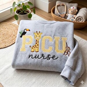 Może przedstawiać: Szara bluza z haftowanym napisem "PICU nurse" czcionką. Litery "PICU" są w żółtą kratkę. Na bluzie wyhaftowano tukana, żyrafę i małpę.