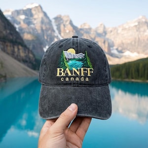 Puede incluir: Gorra de béisbol gris oscuro con el texto bordado "BANFF CANADA". El diseño presenta una escena de montaña con un lago, árboles y un sol. La gorra está hecha de un material suave y desgastado.