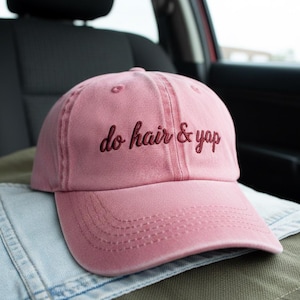Peut inclure: Casquette de baseball rose poudré avec l'inscription brodée "do hair & yap" en rose foncé. Le chapeau est posé sur une veste en jean et une surface verte, avec un siège de voiture en arrière-plan.