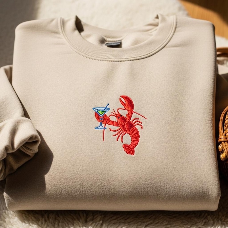 May include: Beige sweatshirt with an embroidered design of a red lobster holding a blue cocktail. The sweatshirt is made of a soft material and has a crew neck. The lobster design is detailed and eye-catching.