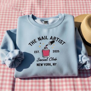 Op de afbeelding: Een lichtblauwe sweatshirt met de tekst "THE NAIL ARTIST Social Club NEW YORK, NY" in zwarte en gouden borduursels. Het ontwerp bevat een nagellakflesje en het jaar 2025. Een strohoed ligt ernaast.