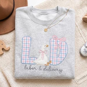 Könnte beinhalten: Ein graumeliertes Sweatshirt mit Rundhalsausschnitt und einer Stickerei der Buchstaben L und D in blau-rosa Karomuster. Eine weiße Gans mit einer rosa Schleife sitzt zwischen den Buchstaben, darunter ist der Text labor & delivery eingestickt.