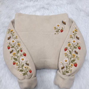 May include: A beige sweatshirt featuring intricate embroidery on both sleeves and the upper right chest. The design displays a botanical pattern of white daisies, yellow wildflowers, red strawberries, and black and yellow bumblebees.
