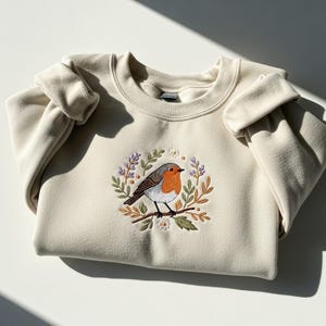 May include: Cream-colored sweatshirt featuring an embroidered robin perched on a branch, surrounded by a wreath of leaves and flowers. The bird has a red breast, brown wings, and a white belly. The design is centered on the chest.