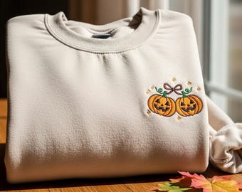 Puede incluir: Sudadera beige con un diseño bordado de dos calabazas sonrientes atadas con un lazo marrón. Las calabazas son naranjas con vides verdes y pequeñas estrellas amarillas. La sudadera está doblada sobre una superficie de madera.