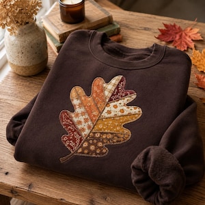 May include: A dark brown crewneck sweatshirt featuring a large, central oak leaf applique. The leaf is constructed from various fabric patches in shades of orange, yellow, and brown, each displaying distinct floral, plaid, and pumpkin patterns.