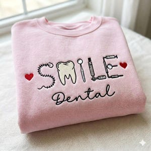 May include: A light pink sweatshirt with the word "SMILE" and "Dental" embroidered in black. The design includes a tooth, dental tools, and red hearts. The sweatshirt is folded.