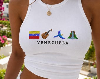 May include: A white sleeveless tank top featuring an embroidered design of the Venezuelan flag, a brown acoustic guitar, a blue and yellow macaw, and a waterfall. The word VENEZUELA is printed in black capital letters below the icons.