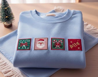 Könnte beinhalten: Hellblaues Sweatshirt mit gestickten Weihnachtsmotiven. Die Aufnäher zeigen einen Weihnachtsmann, Zuckerstangen, ein verpacktes Bonbon und eine Schleife mit einer Schere. Das Sweatshirt ist auf einem hellblauen Tuch gefaltet.
