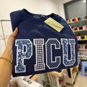 May include: A navy blue sweatshirt featuring the embroidered word "PICU". The letters are decorated with floral, gingham, and polka dot patterns. The sweatshirt has a crew neck and a tag attached.