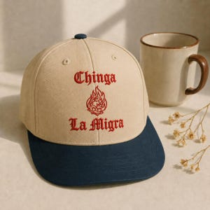 Può includere: Un cappellino da baseball beige con visiera blu scuro. La parte anteriore presenta una scritta ricamata in rosso che recita Chinga La Migra, con un grafico a fiamma ricamato in rosso tra le due righe di testo. Il cappellino ha un bottone in cima.