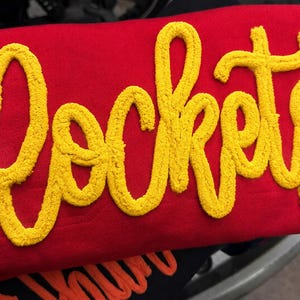 Könnte beinhalten: Ein leuchtend rotes Sweatshirt mit dem Wort Rockets in großen, gelben, strukturierten Schreibschriftbuchstaben im Chenille-Stil auf der Vorderseite. Die gelben Buchstaben haben eine erhabene, flauschige Optik auf dem glatten roten Stoff.