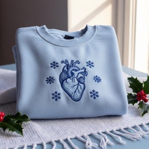 Op de afbeelding: Een lichtblauw sweatshirt met een gedetailleerd blauw anatomisch hartontwerp omringd door blauwe sneeuwvlokken. Het sweatshirt is opgevouwen op een witte deken met franjes, met hulst en rode bessen.