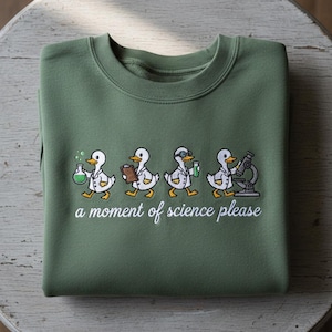 Peut inclure: Un sweat-shirt vert olive avec un motif brodé de quatre canards de dessin animé en blouses de laboratoire, chacun tenant du matériel scientifique. Le texte "a moment of science please" est brodé en dessous.
