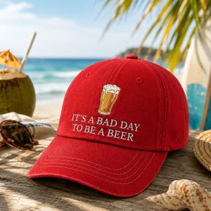 以下が含まれることがあります： 「IT'S A BAD DAY TO BE A BEER」の刺繍とビールグラスのグラフィックが入った赤い野球帽。 海辺の木製の表面に置かれ、ココナッツドリンクとサングラスが背景にあります。