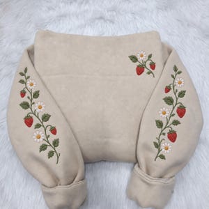 May include: A beige sweatshirt featuring detailed embroidery of strawberry vines with red strawberries, green leaves, and white daisy flowers. The embroidery extends down the length of both sleeves and appears as a small cluster on the upper right.