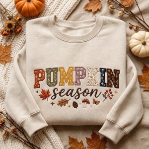 May include: A beige crewneck sweatshirt featuring the word PUMPKIN in large, multi-coloured block letters with floral patterns, and the word season in brown cursive script. Below the text are embroidered autumn motifs including a maple leaf, oak leaf, pinecone, mushroom, butterfly, and berries.