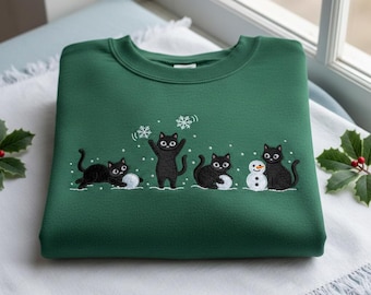 May include: A dark green sweatshirt with an embroidered design of black cats in a snowy scene. The cats are building a snowman and playing with snowflakes. The sweatshirt is folded on a white surface.