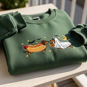 May include: Green sweatshirt featuring embroidered Halloween-themed dachshunds. One is dressed as a hot dog, the other as a ghost holding a pumpkin. Bats, stars, and a crescent moon complete the design. A cozy, festive garment.