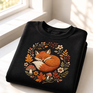 May include: A black sweatshirt featuring a circular embroidered design of a sleeping orange fox curled in the center. The fox is surrounded by a wreath of autumn-themed elements including red and orange maple leaves, small white flowers, and mushrooms.