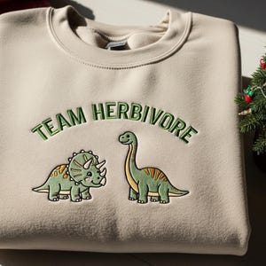 Op de afbeelding: Een beige sweatshirt met de geborduurde tekst "TEAM HERBIVORE" in groen, boven twee cartoon dinosaurussen. Een triceratops en een langnekdinosaurus zijn afgebeeld in groen en geel. De sweatshirt is opgevouwen.