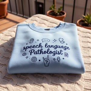 Può includere: Felpa azzurra con la scritta "speech language pathologist" in blu scuro. Il design include icone di orecchio, fumetto, stetoscopio, cervello, mano, microfono e documento. La felpa è piegata su una superficie strutturata color crema.