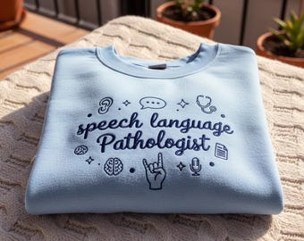 May include: A light blue sweatshirt with the words "speech language pathologist" in dark blue script. The design includes icons of an ear, speech bubble, stethoscope, brain, hand, microphone, and document. The sweatshirt is folded on a cream-colored, textured surface.