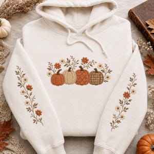 Op de afbeelding: Een crèmekleurige hoodie met een borduursel op de borst van vier pompoenen in verschillende tinten oranje en bruin, omringd door kleine bloemen en bladeren. Bijpassend bloemenborduursel loopt langs beide mouwen naar beneden.